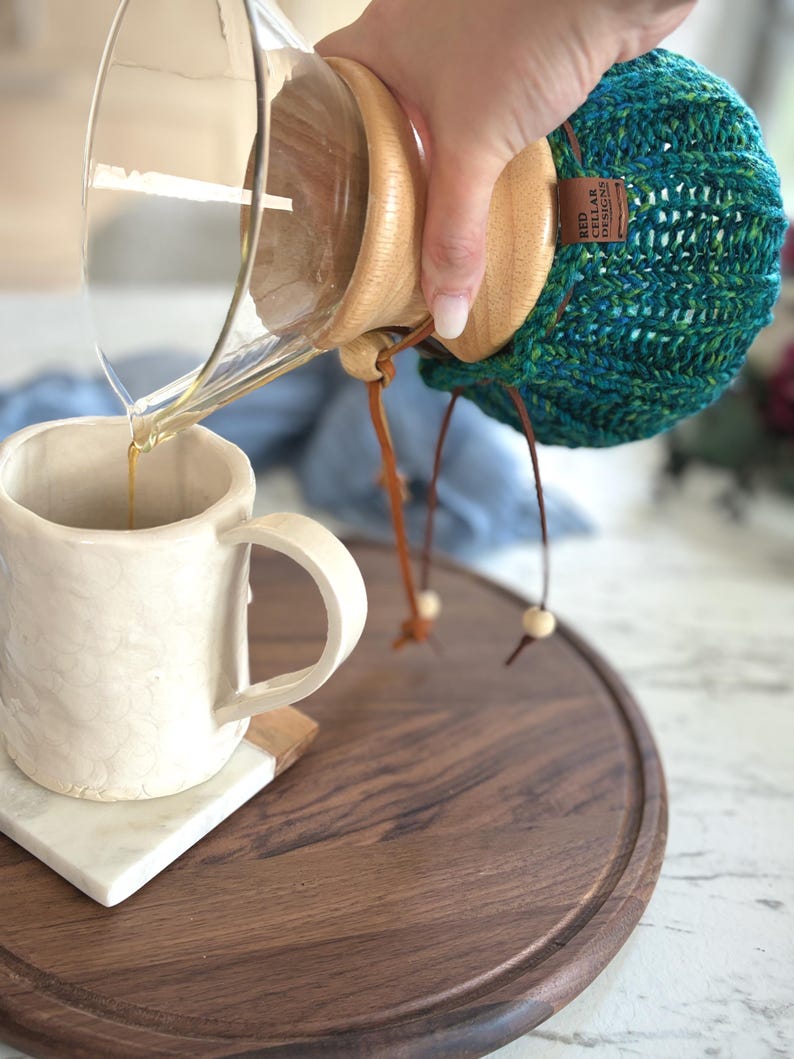 K&ouml;nnte beinhalten: Ein klarer Glas-Kaffeebereiter mit Holzkragen und einem blaugr&uuml;nen Strick&uuml;berzug, der ein braunes Etikett mit der Aufschrift RED CELLAR DESIGNS tr&auml;gt. Der Kaffeebereiter gie&szlig;t Fl&uuml;ssigkeit in eine wei&szlig;e Keramiktasse mit strukturierter Oberfl&auml;che.