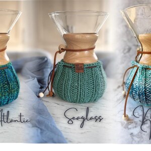 K&ouml;nnte beinhalten: Drei Glas-Kaffeebereiter mit Holzkragen und Lederb&auml;ndern, jeweils mit einem gerippten Strick&uuml;berzug. Die &Uuml;berz&uuml;ge sind beschriftet mit Wild Atlantic in Blaugr&uuml;n, Seaglass in Hellgr&uuml;n und Mourne Mist in blau-wei&szlig; meliertem Garn.