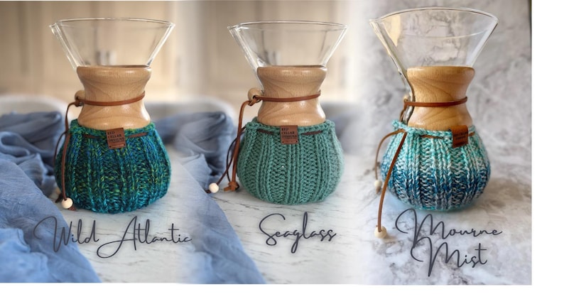 K&ouml;nnte beinhalten: Drei Glas-Kaffeebereiter mit Holzkragen und Lederb&auml;ndern, jeweils mit einem gerippten Strick&uuml;berzug. Die &Uuml;berz&uuml;ge sind beschriftet mit Wild Atlantic in Blaugr&uuml;n, Seaglass in Hellgr&uuml;n und Mourne Mist in blau-wei&szlig; meliertem Garn.