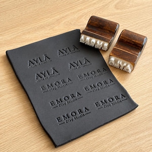 May include: Two wooden stamps with white rubber lettering and a black rectangular sheet featuring embossed impressions of the logos AYLA HOUSE OF CREATION and EMORA Clay Studio. The stamps have dark brown stained wooden handles and white rubber bases.