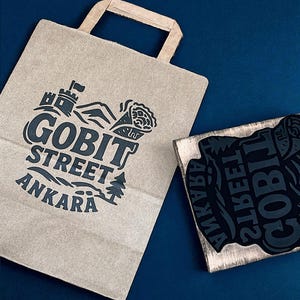 Puede incluir: Una bolsa de papel marrón con asa y un logotipo negro que dice "GOBIT STREET ANKARA" con un castillo, una montaña y un gráfico de árbol. Un sello de madera con el mismo logotipo está al lado de la bolsa.