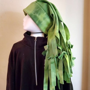 May include: A green fleece hat with a long, pointed shape and dangling strips. The hat has a tie-dye effect with lighter and darker shades of green. The hat is worn over a black zip-up sweatshirt.