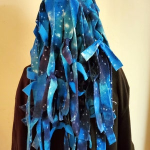 May include: A blue and white galaxy-themed fleece hood with long, knotted strips hanging down. The hood is worn over a dark-colored top, creating a striking contrast. The fleece design resembles a starry night sky.