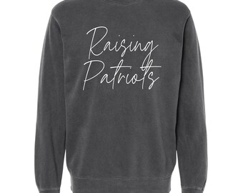 May include: A dark gray sweatshirt with the words "Raising Patriots" in white script across the front. The sweatshirt has a crew neck and long sleeves. The fabric appears to be a soft, comfortable material.