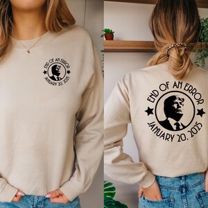 May include: Beige sweatshirt with a black graphic on the front and back. The graphic includes a portrait, the text "END OF AN ERROR" and "JANUARY 20, 2025". The sweatshirt has long sleeves and a crew neck.