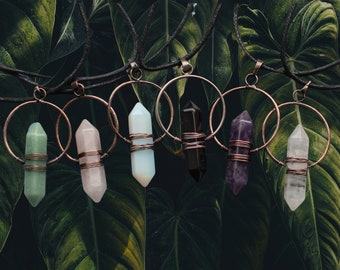 May include: Seven different colored crystal point pendants on copper wire wrapped in copper hoops. The pendants are suspended from black cords.