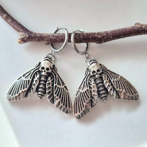 May include: A pair of silver-toned earrings featuring a moth design with a skull at the head. The earrings are suspended from small hoop closures and are displayed on a brown twig against a white background. The moth wings have detailed patterns.
