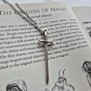 May include: A silver sword pendant necklace with a chain. The pendant is a simple, flat sword design with a pointed tip. The necklace is lying on a book with the text "The Origins of Magic" visible.