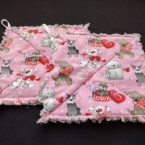 May include: Two pink quilted pot holders with a frayed edge. The fabric features a pattern of dogs holding hearts and the text "XOXO".