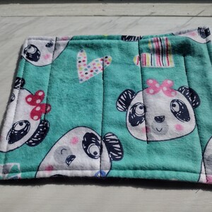 May include: A square, teal fabric item with a white border, featuring a repeating pattern of panda faces with pink bows. The design includes a heart and striped details. The item is likely a coaster or small mat.