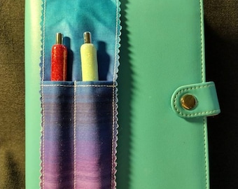 May include: A teal planner with a fabric pen holder attached. The pen holder has a gradient design of blue, purple, and pink. Two pens are visible: one red and one light green. The planner has a snap closure.