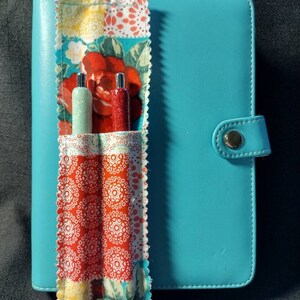 May include: A turquoise planner with a fabric pen holder attached. The pen holder has a patchwork design with red, white, and green floral patterns. Two pens are inserted into the holder.