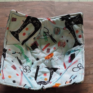 May include: A blue quilted hot pad with a sewing themed print. The print features black sewing machines, scissors, spools of thread, buttons, and other sewing supplies.
