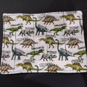 May include: White fabric placemat with a dinosaur theme. The placemat features various dinosaurs, including Tyrannosaurus Rex, Stegosaurus, Triceratops, Brachiosaurus, and Spinosaurus. The dinosaurs are depicted in green, brown, and yellow colors.