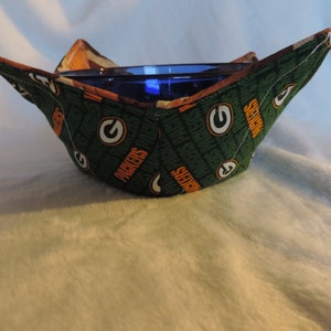 May include: Green and orange fabric bowl cozy with the Green Bay Packers logo and the words "Packers" and "Green Bay" repeated. The cozy is holding a blue glass bowl.