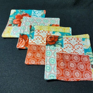 May include: Four colorful fabric coasters with a patchwork design featuring red, yellow, blue, and white floral patterns. The coasters are made of a soft, quilted fabric and have a decorative stitched edge.