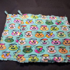 May include: A square quilted potholder featuring a repeating pattern of colorful teacups on a light blue background. Each teacup is decorated with a dark heart shape in the center. The edges of the fabric are finished with a fringed, knotted border.