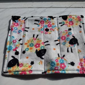 May include: A white, rectangular fabric item with a sheep and floral pattern. The sheep are black with floral wool, and the flowers are pink, blue, yellow, and orange. The item is likely a coaster or small mat.