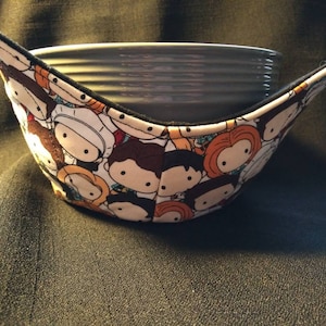 May include: A white fabric bowl cozy with a black trim and a colorful cartoon print of characters. The cozy is holding a stack of gray ceramic bowls.