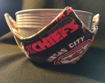 May include: A black bowl cozy featuring the Kansas City Chiefs logo in red and white. The cozy is wrapped around a gray bowl. The text "CHIEFS" and "KANSAS CITY" are visible on the fabric.