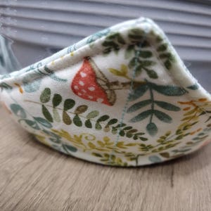 May include: A white fabric bowl cozy with a botanical print featuring green leaves, yellow accents, and a red mushroom. The cozy is designed to hold a bowl and protect hands from heat.