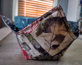 May include: A fabric bowl cozy with a floral and bird print design. The cozy is black and white with a beige background and features a red rose and a brown bird.