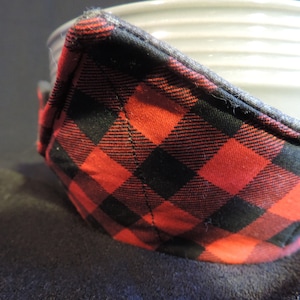 May include: Close-up of a red and black plaid fabric bowl cozy. The cozy is made of a soft, quilted fabric and has a gray trim.