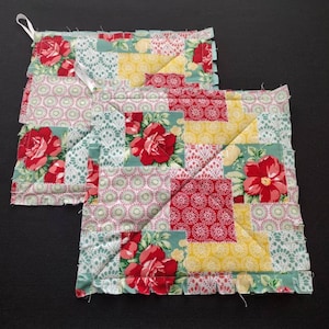 May include: Two quilted pot holders with a patchwork design featuring red roses, green leaves, and yellow, red, and white geometric patterns.