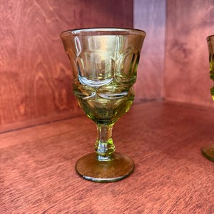 May include: A vintage green glass cordial glass with a detailed pattern on the bowl and a delicate stem. The glass has a gold rim and sits on a round base. The glass is displayed on a wooden surface.