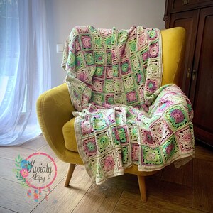 May include: A colourful crochet blanket, draped over a yellow armchair. The blanket features a square pattern with pink, green, and cream-coloured squares. The blanket's design is intricate, with a floral motif in each square. The chair has wooden legs.