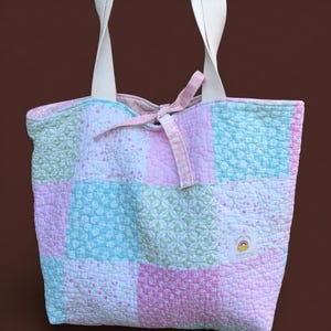 May include: A patchwork tote bag in pastel shades of pink, blue, and green. It has white straps and a pink ribbon tie. The bag is quilted and features a small label with a rainbow design.