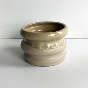 Op de afbeelding: Een kleine, keramische plantenpot met een lichtbruine glazuur aan de bovenkant en een witte glazuur aan de onderkant. De pot heeft een afgeronde vorm en een licht uitlopende rand.