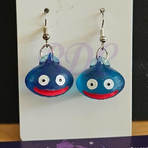 Op de afbeelding: Een paar blauwe hars oorbellen in de vorm van lachende cartoon slimes. Elke oorbel heeft witte ogen en een rode mond, hangend aan zilverkleurige oorhaakjes.
