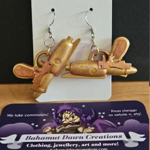 May include: A pair of gold-colored earrings shaped like ray guns. The earrings have a metallic finish and are suspended from silver-colored fish hook ear wires. The earrings are displayed on a white card with a business card below.