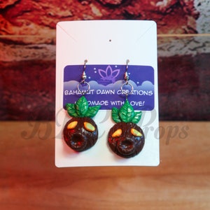 Puede incluir: Un par de pendientes con una escultura de arcilla marrón y verde de un personaje con una expresión de sorpresa. Los pendientes están en una tarjeta blanca con el texto "Bahamut Dawn Creations" y "Made with Love!"