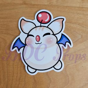 Puede incluir: Una pegatina troquelada con un personaje de dibujos animados con cuerpo blanco, orejas grandes y pequeñas alas azules. El personaje tiene un orbe rojo en la cabeza y mejillas rosadas, con una expresión feliz. La pegatina está sobre una superficie de madera.