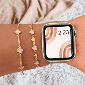 May include: A gold smartwatch with a white watch face featuring a watercolor rainbow design and the time 2:23. The watch is worn on a wrist with two gold bracelets, one with clover charms and the other with heart charms.