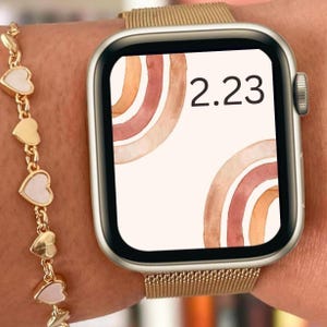 May include: A gold Apple Watch with a gold metal band. The watch face is white with two watercolor-style rainbows in shades of brown, orange, and pink. The time is 2:23.