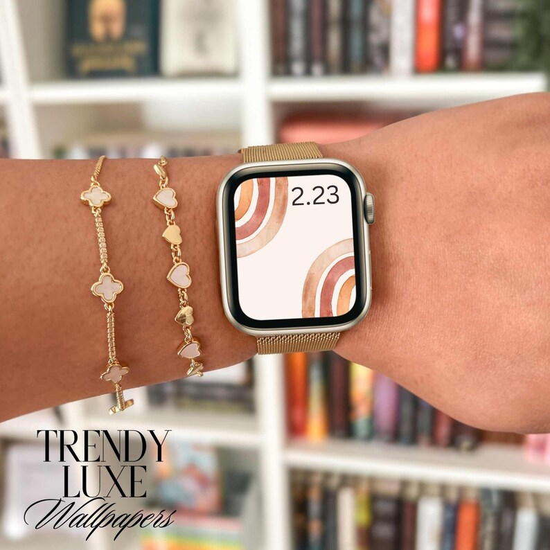 May include: A gold Apple Watch with a white watch face featuring a watercolor design of abstract arches in shades of brown, orange, and pink. The time is displayed as 2.23. The watch is worn on a person's wrist with two gold bracelets.