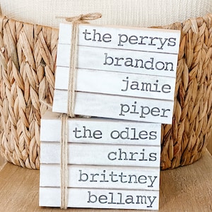 May include: Stacked white wooden blocks with black text, reading "the Perrys, Brandon, Jamie, Piper, the Odles, Chris, Brittney, Bellamy".