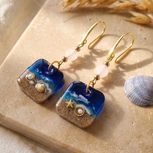 Könnte beinhalten: Ein Paar Ohrhänger mit quadratischen Harzanhängern, die eine Strandszene mit blauem Meerwasser, weißem Schaum und beigem Sand zeigen. Jeder Ohrring ist mit einem kleinen goldenen Seestern, einer winzigen Perle und einer blassrosa Perle an einem Goldhaken verziert.