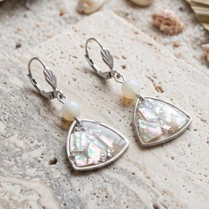 Peut inclure: Une paire de boucles d'oreilles à fermoir levier argenté avec un pendentif triangulaire rempli de fragments de coquillages irisés. Chaque boucle d'oreille comporte une perle blanche ronde placée au-dessus de l'incrustation triangulaire.
