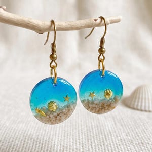 Könnte beinhalten: Ein Paar runde Ohrhänger mit einem blauen Harzverlauf, der in eine sandfarbene Basis übergeht. Jeder Ohrring enthält eingebettete goldfarbene Miniatur-Muscheln und Seestern-Anhänger, befestigt an goldfarbenen Metall-Ohrhaken.