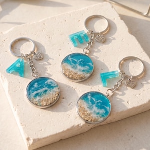 Peut inclure: Trois porte-clés circulaires présentant une scène d'océan en résine avec de l'eau bleue, de l'écume blanche et du sable beige. Chaque porte-clés comprend un anneau en métal argenté, un petit cœur avec le texte handmade with love et une lettre bleue.