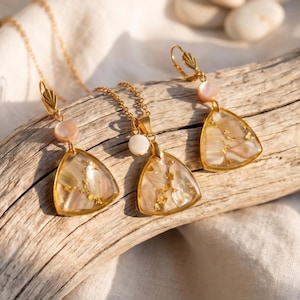 Op de afbeelding: Een goudkleurige sieradenset met een ketting en oorbellen. Elk stuk heeft een driehoekige hanger met een iriserende, parelmoer afwerking en goudblad accenten. De oorbellen en ketting zijn versierd met een roze en witte kraal.