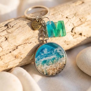 Peut inclure: Un porte-clés argenté avec un pendentif rond représentant une scène de plage, une lettre « M » turquoise et verte, et un charm en forme de cœur avec l'inscription « Made with Love ». La scène de plage montre des vagues bleues et blanches et du sable.