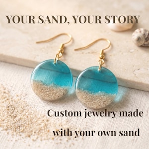 Peut inclure: Une paire de boucles d'oreilles pendantes rondes avec un crochet doré et un pendentif circulaire en résine bleue transparente sur une base de sable. Le texte indique : YOUR SAND, YOUR STORY. Bijoux personnalisés faits avec votre propre sable.