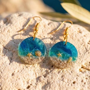 Può includere: Un paio di orecchini pendenti rotondi con un design in resina che imita una scena di spiaggia con un gradiente di oceano blu, sabbia bianca e piccoli ciondoli dorati a forma di stella marina e conchiglia, fissati a ganci in metallo dorato.