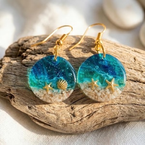 May include: A pair of round drop earrings featuring a resin ocean scene with a gradient of deep blue to turquoise water, embedded with tiny golden starfish and a seashell, resting on a layer of white sand, attached to gold-toned hooks.