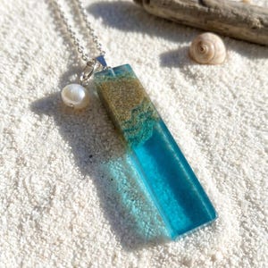 Puede incluir: Un collar plateado con un colgante rectangular que representa una escena de playa. El colgante tiene una capa de arena, resina azul y un dije de perla. Una concha y madera flotante están en el fondo.